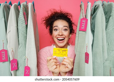 Young Surprised Smiling Female Costumer Woman 20s Wear Sweater Stand Near Clothes Rack With Tag Sale In Store Showroom Hold In Hand Credit Bank Card Isolated On Plain Pink Background Studio Portrait