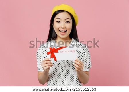 Young surprised woman of Asian ethnicity in white polka dot t-shirt yellow beret hold gift certificate coupon voucher card for store isolated on plain pastel pink background. People lifestyle concept