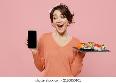 Young surprised woman in sweater hold makizushi sushi roll served on black plate traditional japanese food use mobile cell phone blank screen workspace area isolated on plain pastel pink background.