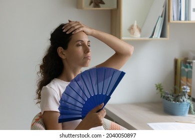 Young sweaty thirsty woman sit indoor at desk feels overheated due unbearable summer hot day, holding handheld blue fan cooling herself. Apartment without air-conditioner, need climate control concept