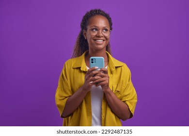 Young sweet dreamy African American woman looks up with smile and holds phone flirting with boyfriend in online chat and exchanging romantic messages on dating apps stands on lilac background. - Powered by Shutterstock