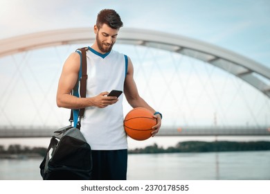 Young tall sportsman walking with sports bag while using mobile phone and holding a basketball. Handsome male basketball player with ball and sports bag holding and looking at mobile phone. - Powered by Shutterstock