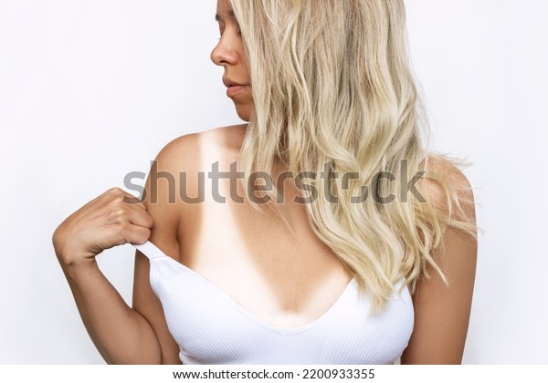 Young tanned blonde woman showing off her tan by\
lifting up strap of swimsuit isolated on white background. Result\
of tanning before and after. Tanned skin, contrast of skin colors.\
Traces of swimwear