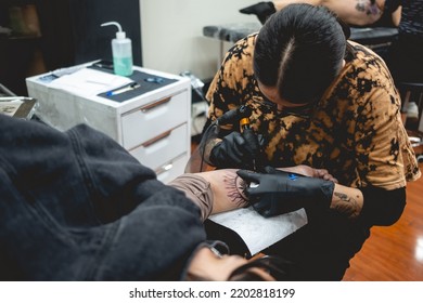 Young Tattoo Artist Girl With Glasses And Mask Making A Tattoo Of 'La Santa Muerte' (Our Lady Of Holy Death) In The Arm Of A Woman With Tattoo Machine