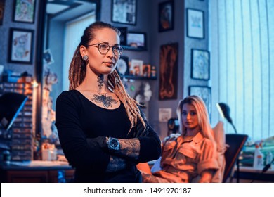 Young tattoo master is standing crossed her hands while attractive girl is waiting her on the couch.