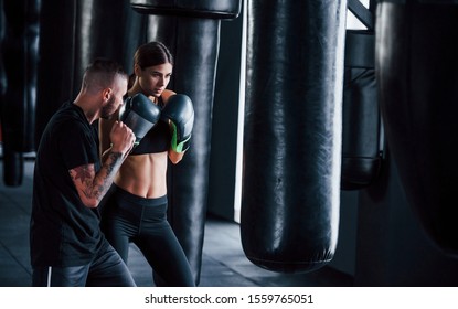 Young Tattooed Boxing Coach Teaches Young Woman In The Gym.