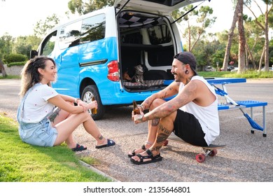 Young tattooed couple drinking beer bottles sitting near by the van with their dogs, longboard and picnic table.
