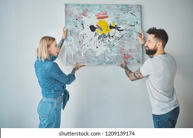 Young Tattooed Couple Hanging Painting On Wall Together At New Apartment