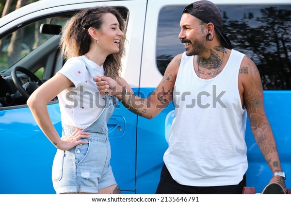 Young tattooed couple laughing and\
looking each other at the side of the van with a\
longboard.