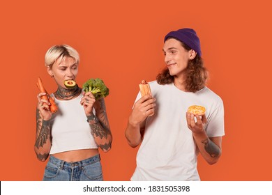 Young Tattooed Diverse Couple Standing Isolated On Orange Background Girlfriend Holding Fresh Vegetables Maintaining Healthy Lifestyle Vs Boyfriend Holding Hot Dog And Donut Junk Food Looking At Each