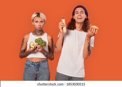 Young Tattooed Diverse Couple Standing Isolated On Orange Background Girlfriend Holding Fresh Vegetables Maintaining Healthy Lifestyle Vs Boyfriend Holding Hot Dog And Donut Junk Food Looking Camera