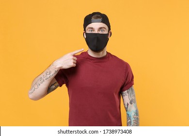 Young Tattooed Man Guy In Casual T-shirt Cap Posing Isolated On Yellow Wall Background Studio Portrait. People Emotions Lifestyle Concept. Mock Up Copy Space. Pointing Index Finger On Black Face Mask