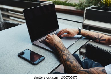 Young tattooed man working on laptop in cafe. Back view of human hands with tattoo using laptop at coworking office desk. Handsome guy typing text on laptop with blank copy space screen mockup