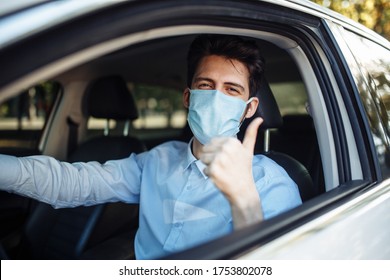 Young Taxi Driver Shows Thumb Up Sign Sitting In The Car And Wearing Protective Sterile Medical Mask, Works Hard During Coronavirus Outbreak. Social Distance, Virus Spread Prevention, Treat Concept.