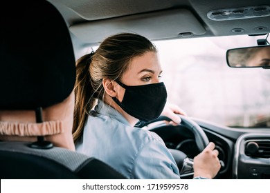 A young taxi driver woman in a black medical mask with blond hair and a turquoise jacket is driving a right-hand drive car. A woman looking at road. Masks protective. Quarantine. Virus. Pandemic.