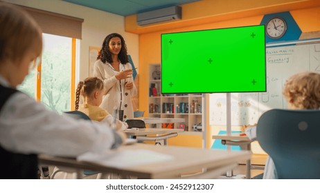 Young Teacher Explaining a Class Assignment to Elementary School Kids on a TV Screen with Green Mock Up Chroma Key. Diverse International Kids Receiving Modern Primary Education