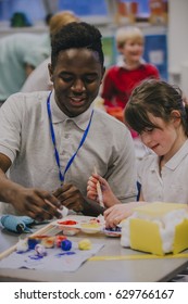 Young Teacher Is Working With One Of His Primary School Students During An Art And Crafts Lesson. They Are Painting And Discussing The Project.