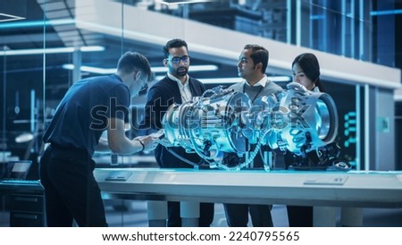 Young Team of Engineers, Project Manager and Machinery Operators Collaborate on a New Type of a Gas Turbine Engine, Standing with Tablet and Laptop Computers in Scientific Technology Lab. High Angle.