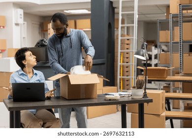 Young team of workers processing packages on a laptop in a busy shipping area, handling custom orders and shipment logistics with taped boxes. Small business local brand environment.