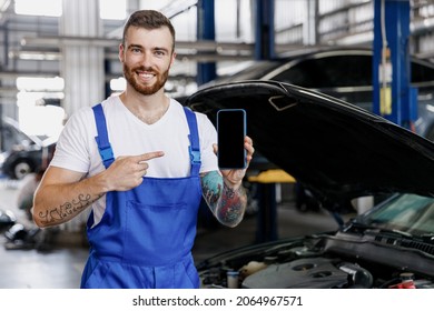 Young Technician Car Mechanic Man In Overalls White T-shirt Use Hold Point Finger On Mobile Cell Phone Blank Screen Area Fix Problem With Raised Hood Bonnet Work In Vehicle Repair Shop Workshop Indoor