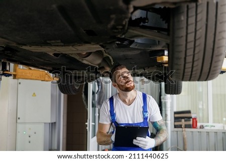 Young technician mechanic man wears denim overalls use hold clipboard papers document writing estimate outlay stand near car lift check technical condition work in vehicle repair shop workshop indoor