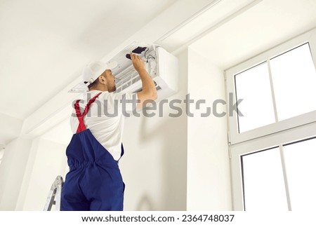 Young technician uses screwdriver and opens up air conditioner unit on white wall at home or in office. Back view, backside shot from behind. AC installation, maintenance and repair service concept