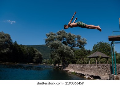 Young Teen Boy Jumping Flying And Diving In The River. Clear Blue Sky And Trees In Distance As A Natural Background.