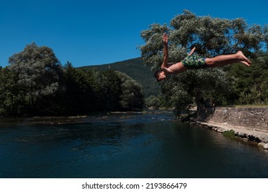 Young Teen Boy Jumping Flying And Diving In The River. Clear Blue Sky And Trees In Distance As A Natural Background.