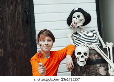 Young teen boy and painted face spider at all hallows eve photo session enjoy pumpkin jack lantern lollipop indoor  Stylish child in orange clothes celebrates halloween near human pirate skeleton