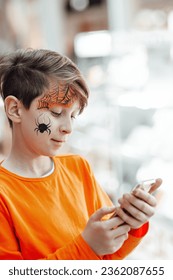 Young teen boy and painted face spider all hallows eve using wireless technology mobile device chatting  Stylish child in orange clothes celebrates halloween holiday enjoy texting sms by phone