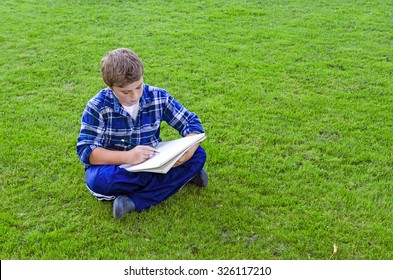 Young Teen Boy With Sketch Pad Drawing  Or Writing While Sitting On Grass Outdoors, With Copy Space