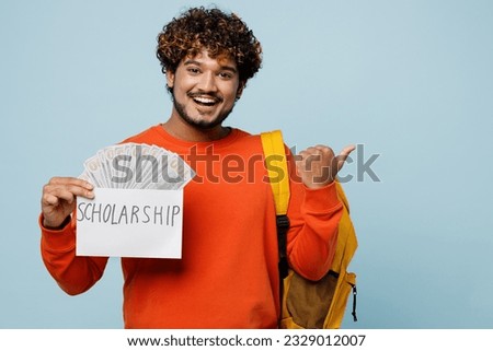 Young teen boy student wears casual clothes backpack bag hold fan of cash money in dollar banknotes, sholarship point aside isolated on plain blue background. High school university college concept
