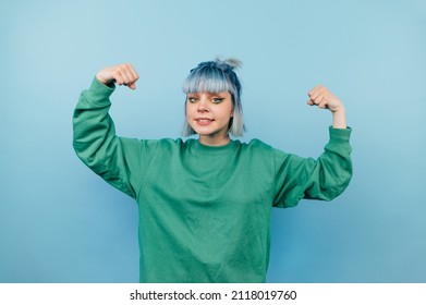 Young teen girl with blue hair shows biceps to the camera and looks with a serious face on a blue background