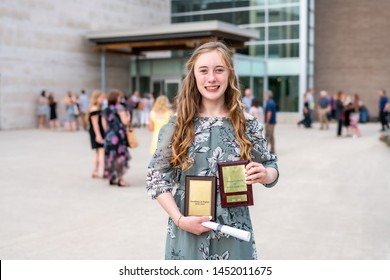 Young Teen Girl/Middle School Student Standing In Front Of School With Awards And Diploma After Grade 8/Middle School Graduation Ceremony With Blurred People In Background.