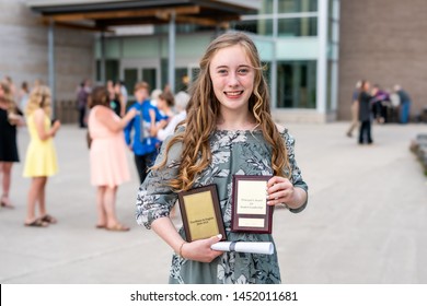 Young Teen Girl/Middle School Student Standing In Front Of School With Awards And Diploma After Grade 8/Middle School Graduation Ceremony With Blurred People In Background.