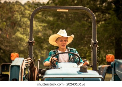 A young teenage boy driving a tractor with a cowboy hat on- A cowboy on a tractor doing farm work- A ranch hand with his hands on the steering wheel of a blue tractor driving it