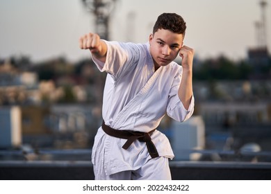 Young teenage boy karate practitioner in white kimono training on the roof top at sunset