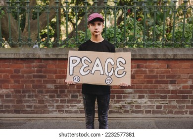 Young teenage boy looking at the camera while holding a peace poster. Young peace activist protesting against war and violence. - Shutterstock ID 2232577381