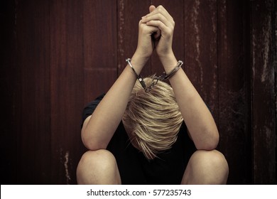Young teenage boy wearing handcuffs sitting with his head bowed between his legs in a concept of crime and capture or abuse