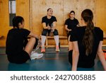 Young teenage female multicultural volleyball players are sitting on indoor court and taking a break after their training. Young sportswomen sitting on volleyball court, refreshing themselves on break