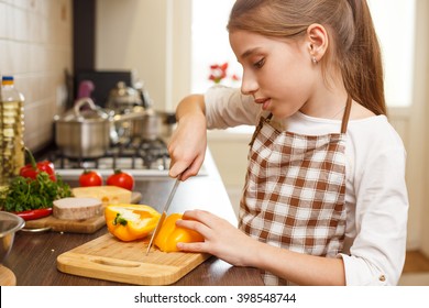 Young teenage girl in apron cutting sweet pepper on board at the kitchen. 