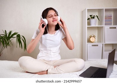 A Young Teenage Girl With Headphones Listening To Music With Her Eyes Closed. The Girl Is Sitting On The Bed In The Room In Comfortable Clothes. Mock Up.