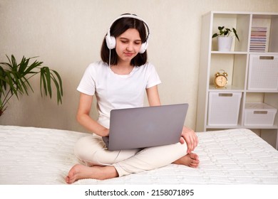 A Young Teenage Girl With Headphones Listening To Music, Working On A Laptop. The Girl Is Sitting On The Bed In The Room In Comfortable Clothes. Mock Up.