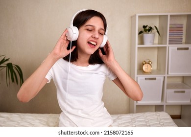 A Young Teenage Girl With Headphones Listens To Music And Sings With Her Eyes Closed. The Girl Is Sitting On The Bed In The Room In Comfortable Clothes. Mock Up.