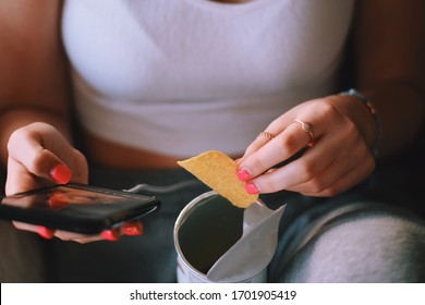 Young Teenage Girl Is Sitting On A Couch At Home Using Mobile Phone And Eating Potato Chips