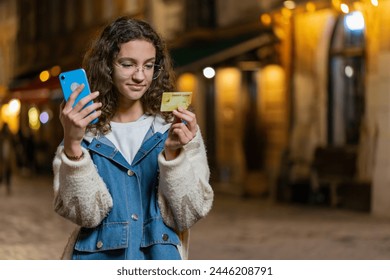 Young teenage girl using credit bank card smartphone while transferring money, purchases online shopping, order food delivery, booking hotel room. Happy woman in urban city street at night. Copy-space - Powered by Shutterstock