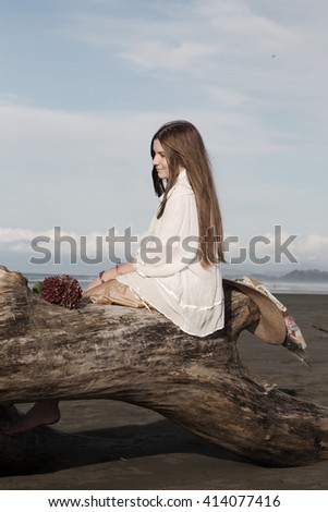 A young, teenage girl wearing a flowing skirt and blouse sitting on driftwood enjoying the late afternoon sun at the beach. Dried flowers and a straw hat are the finishing touches. Vertical format.