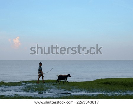 Young teenage goat keeper walking with his domestic pets near a river, people relaxing in a riverbank meadow 