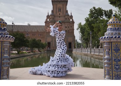 Young Teenage Woman In White Dance Suit With Black Polka Dots And Green Carnations In Her Hair, Dancing Flamenco By A Lake. Flamenco Concept, Dance, Art, Typical Spanish Dance.