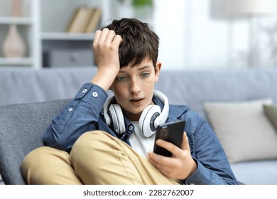 Young teenager boy reading bad news on phone, son sitting on sofa in living room with headphones at home closeup. - Powered by Shutterstock